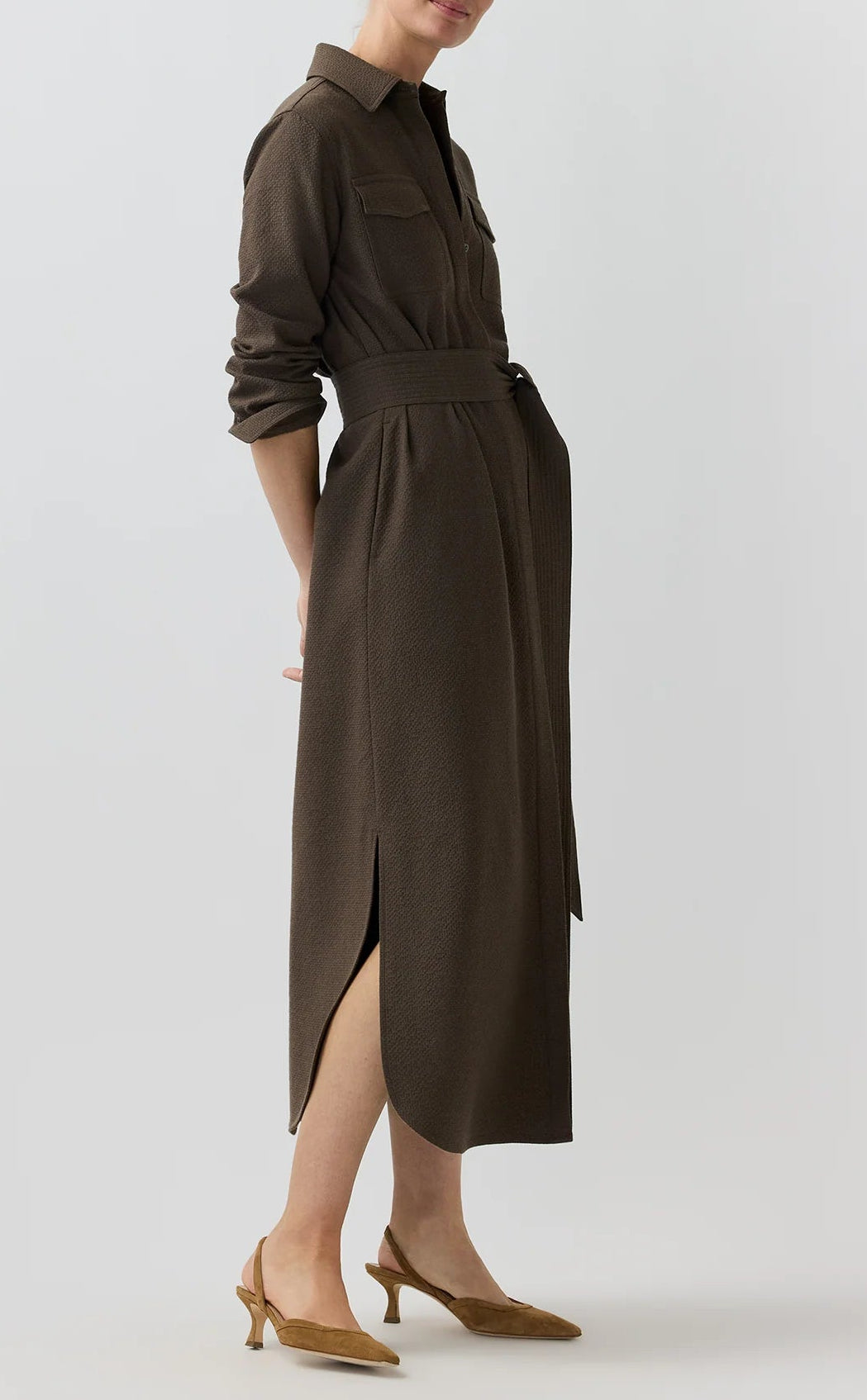 A woman stands against a plain light background wearing the Ann Mashburn Maude Crinkle Cotton/Wool Dress—a long-sleeved, belted brown shirt dress featuring a textured finish, front pockets, and side slits—paired with tan slingback heels.