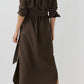 A person stands with their back to the camera, wearing the Ann Mashburn Maude Crinkle Cotton/Wool Dress in brown, styled with beige heeled sandals against a plain light background.