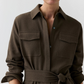 A person wearing the Ann Mashburn Maude Crinkle Cotton/Wool Dress—a textured brown collared shirt dress featuring long sleeves, chest pockets, and a tied waist belt—stands against a plain background.