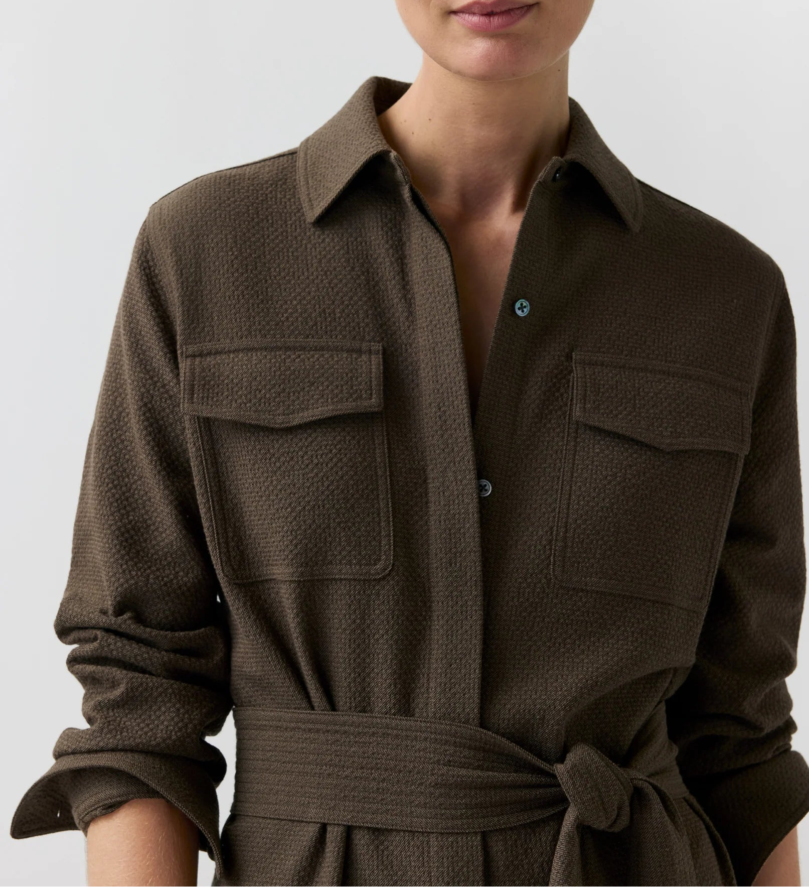 A person wearing the Ann Mashburn Maude Crinkle Cotton/Wool Dress—a textured brown collared shirt dress featuring long sleeves, chest pockets, and a tied waist belt—stands against a plain background.