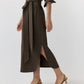 A woman walks against a plain background wearing the Ann Mashburn Maude Crinkle Cotton/Wool Dress—a long brown belted Italian-fabric dress with a side slit—paired with brown low-heeled slingback shoes.