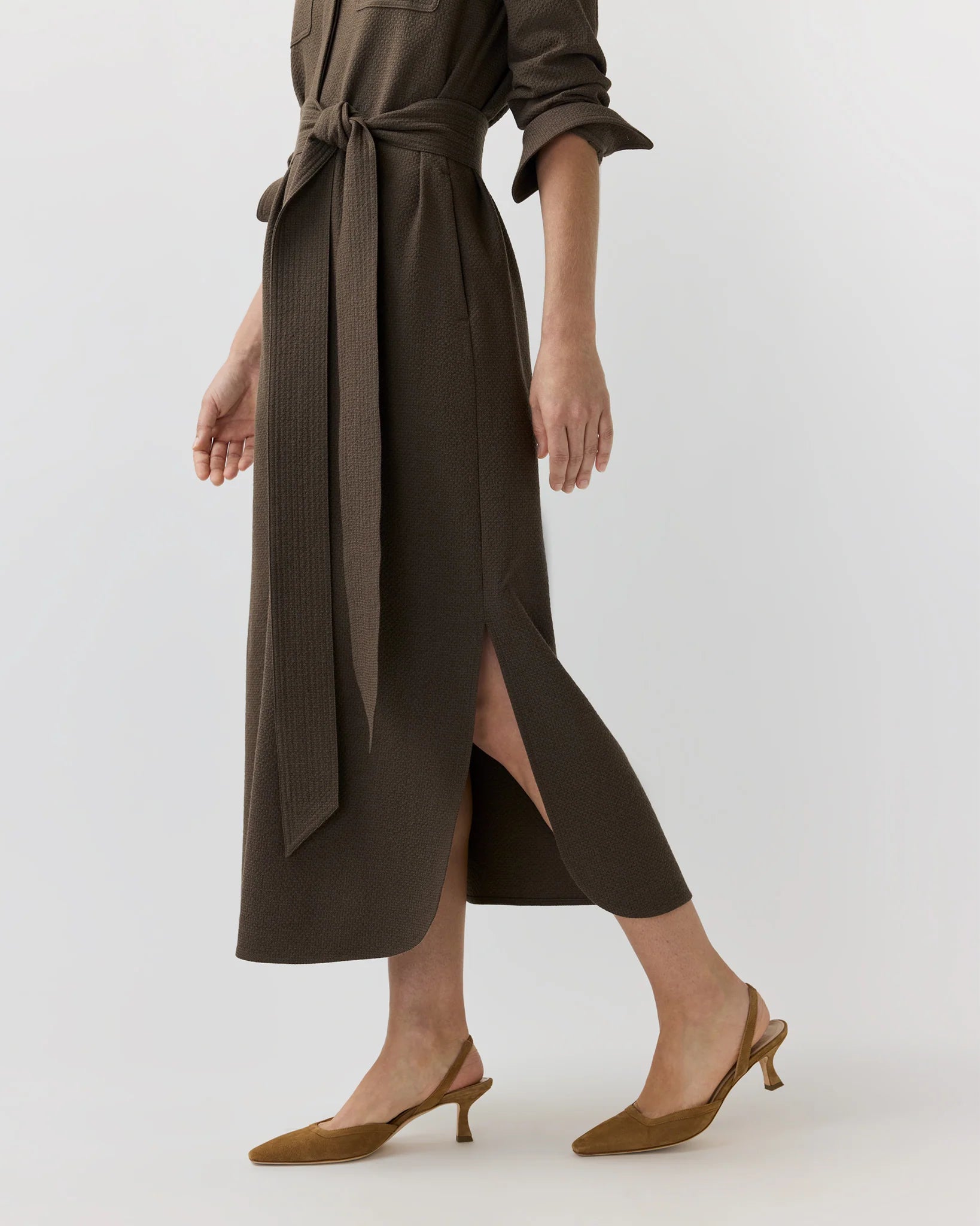 A woman walks against a plain background wearing the Ann Mashburn Maude Crinkle Cotton/Wool Dress—a long brown belted Italian-fabric dress with a side slit—paired with brown low-heeled slingback shoes.