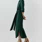 A person stands sideways wearing the dark green Piper Silk Crepe Maxi Dress in crepe de chine with a waist tie and side slits, paired with tan strappy heels.