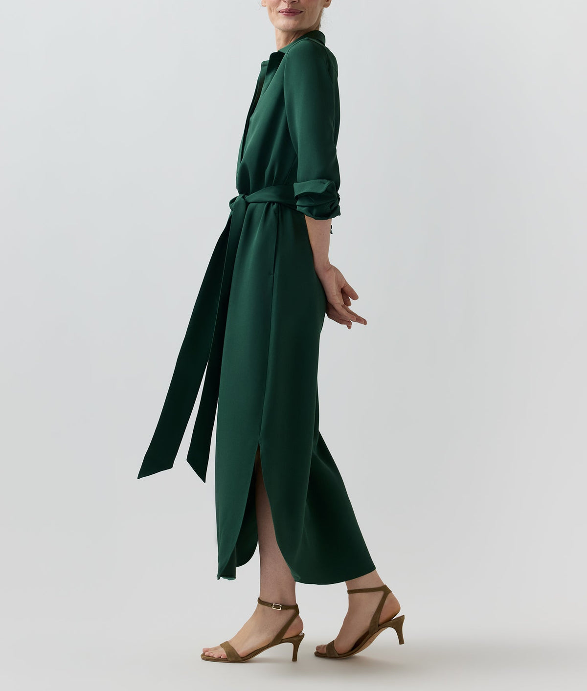 A person stands sideways wearing the dark green Piper Silk Crepe Maxi Dress in crepe de chine with a waist tie and side slits, paired with tan strappy heels.