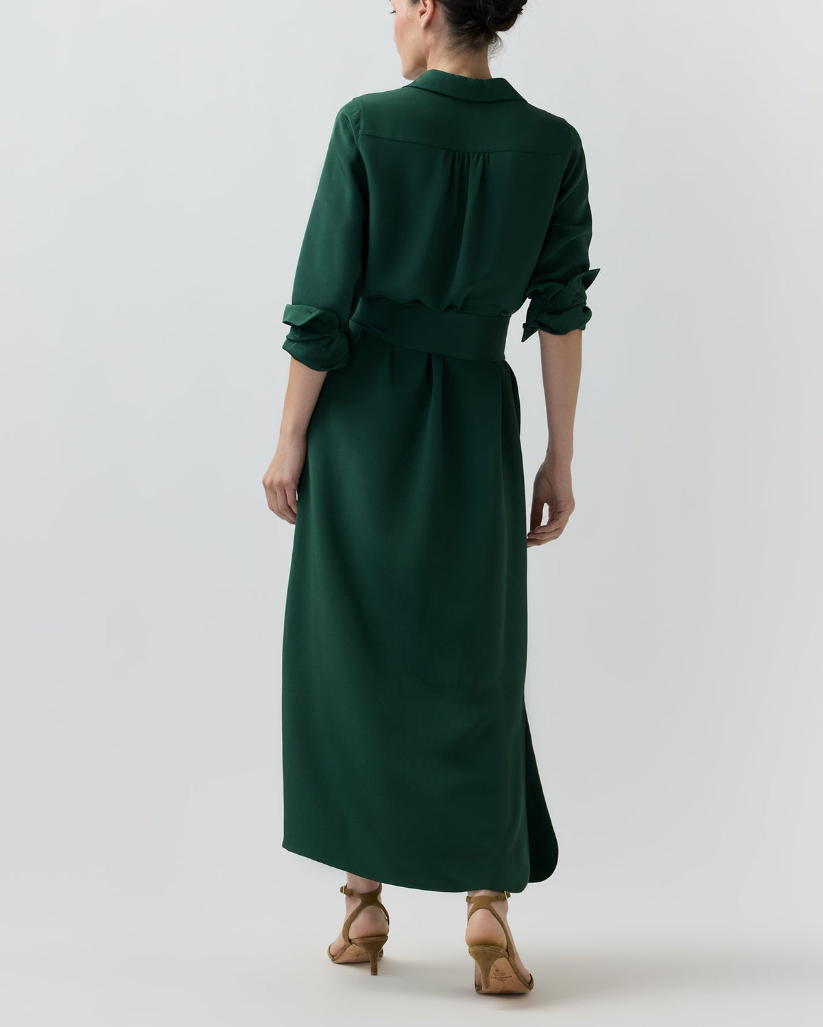 Back view of a person wearing a dark green Piper Silk Crepe Maxi Dress with a collar and waist tie, paired with tan heeled sandals.