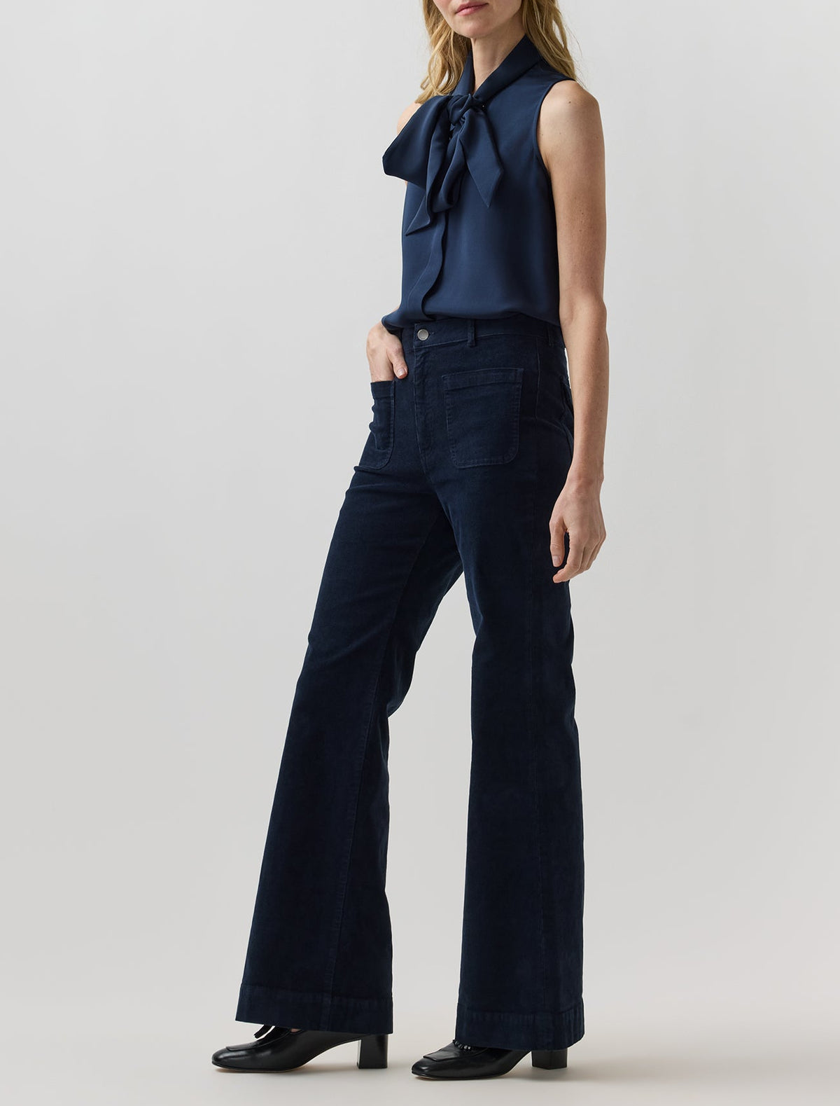 A person wears a sleeveless navy blue blouse with a bow tie neck and Ann Mashburn Camille Patch-Pocket Stretch Corduroy Jean, which are dark blue, wide-leg, and feature front patch pockets.