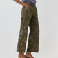 Wearing the Ann Mashburn Frankie Stretch Twill Cropped Pant in olive green camo, featuring a minimal waistband, styled with a blue and white gingham shirt and tan heels, this look exudes effortless style with hands casually in pockets against a plain backdrop.