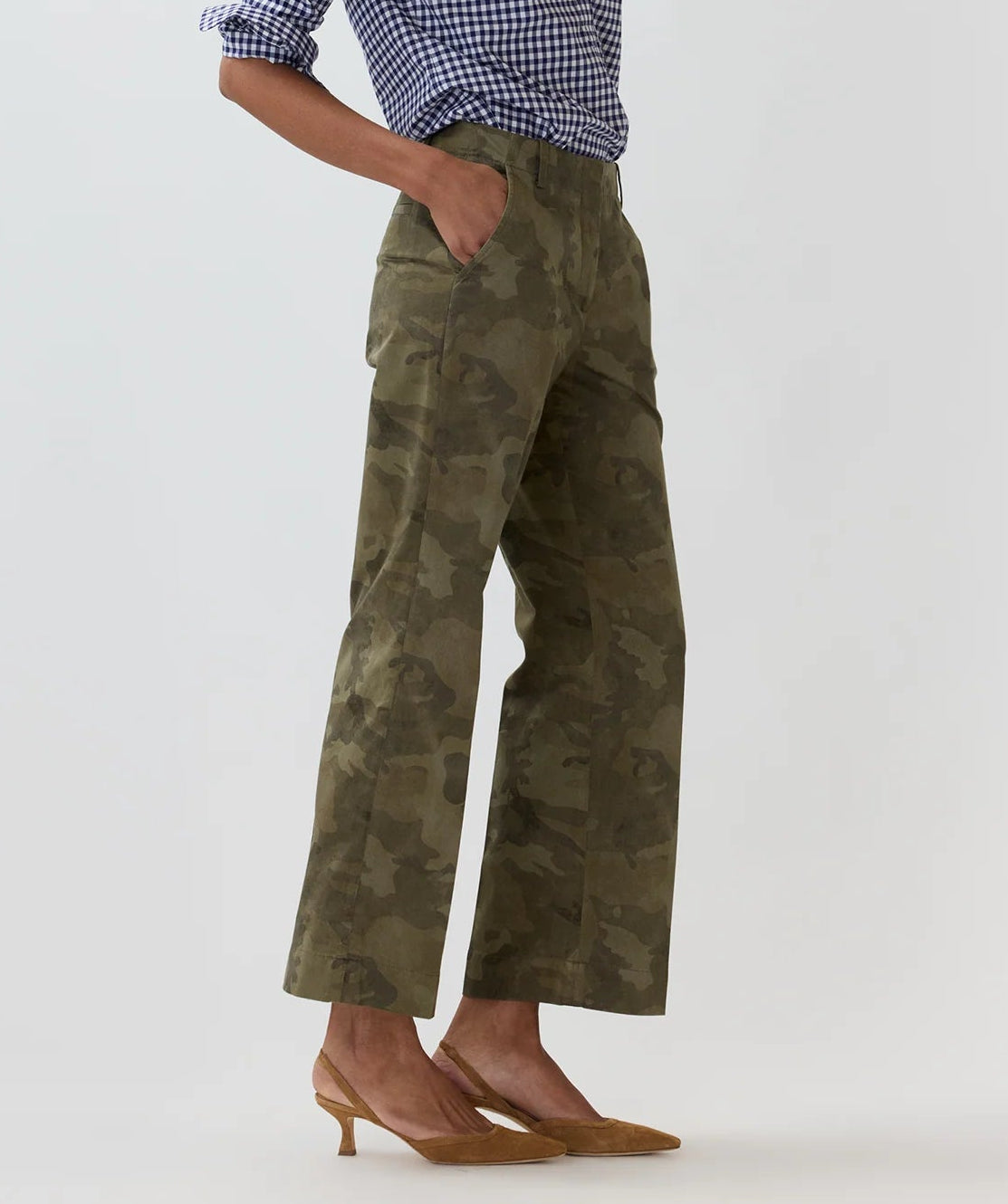 Wearing the Ann Mashburn Frankie Stretch Twill Cropped Pant in olive green camo, featuring a minimal waistband, styled with a blue and white gingham shirt and tan heels, this look exudes effortless style with hands casually in pockets against a plain backdrop.