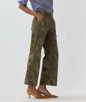 Wearing the Ann Mashburn Frankie Stretch Twill Cropped Pant in olive green camo, featuring a minimal waistband, styled with a blue and white gingham shirt and tan heels, this look exudes effortless style with hands casually in pockets against a plain backdrop.