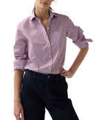 Wearing the Sidney Banker Stripe Poplin Shirt by Ann Mashburn—a red and white menswear stripe button-up crafted from Italian poplin with a spread collar—this person has the sleeves rolled up and pairs it with dark pants, standing with one hand on hip.