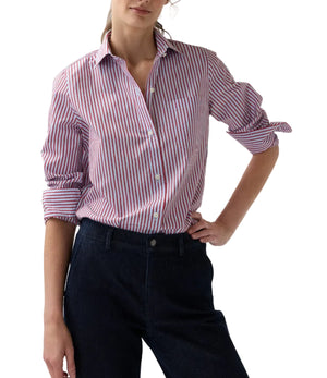 Wearing the Sidney Banker Stripe Poplin Shirt by Ann Mashburn—a red and white menswear stripe button-up crafted from Italian poplin with a spread collar—this person has the sleeves rolled up and pairs it with dark pants, standing with one hand on hip.