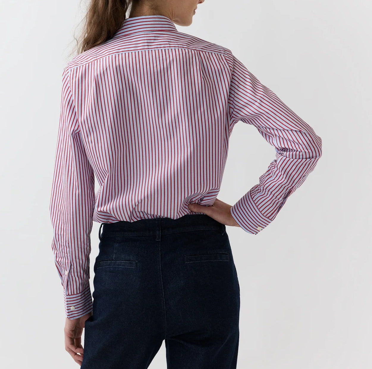 A person stands with their back to the camera, wearing the Ann Mashburn Sidney Banker Stripe Poplin Shirt—a red and white menswear stripe button-down crafted from fine Italian poplin—paired with dark blue jeans, set against a plain light background.