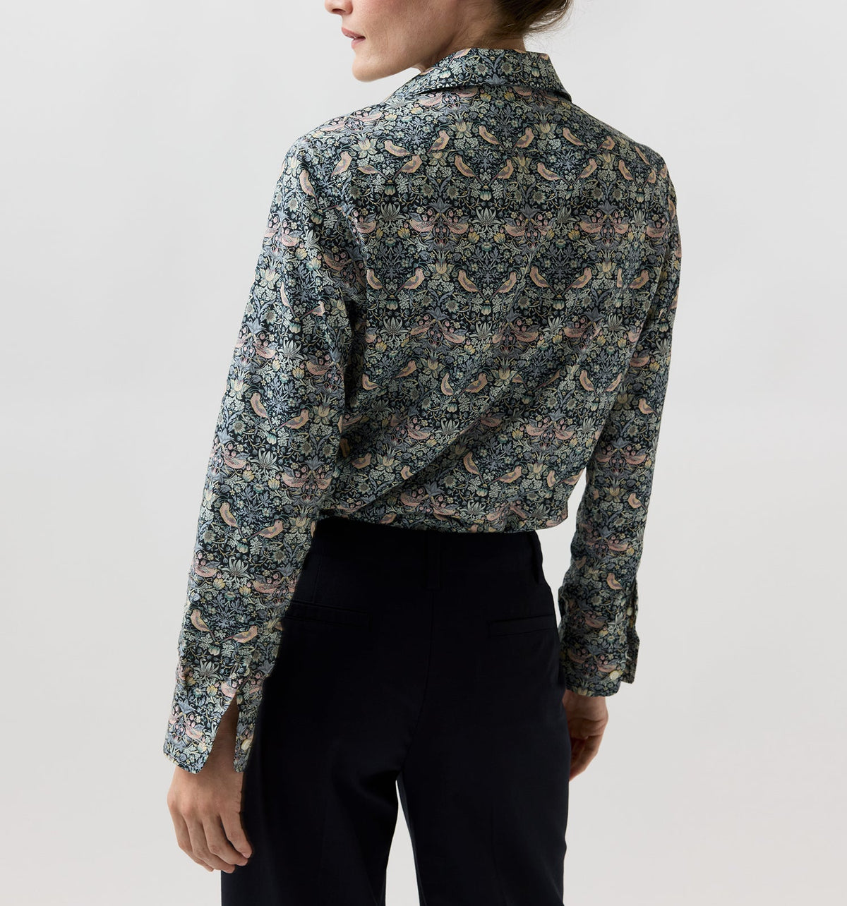 Back view of a person in an Ann Mashburn Tomboy Popover Liberty Shirt with a multicolored floral and bird pattern, tucked into black trousers.