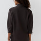 A person with curly hair stands with their back to the camera against a plain light background, wearing the Ann Mashburn Violetta Bi-Stretch Tricotine Tunic—a modern, dark, long-sleeve, stretchy top—paired with matching pants.