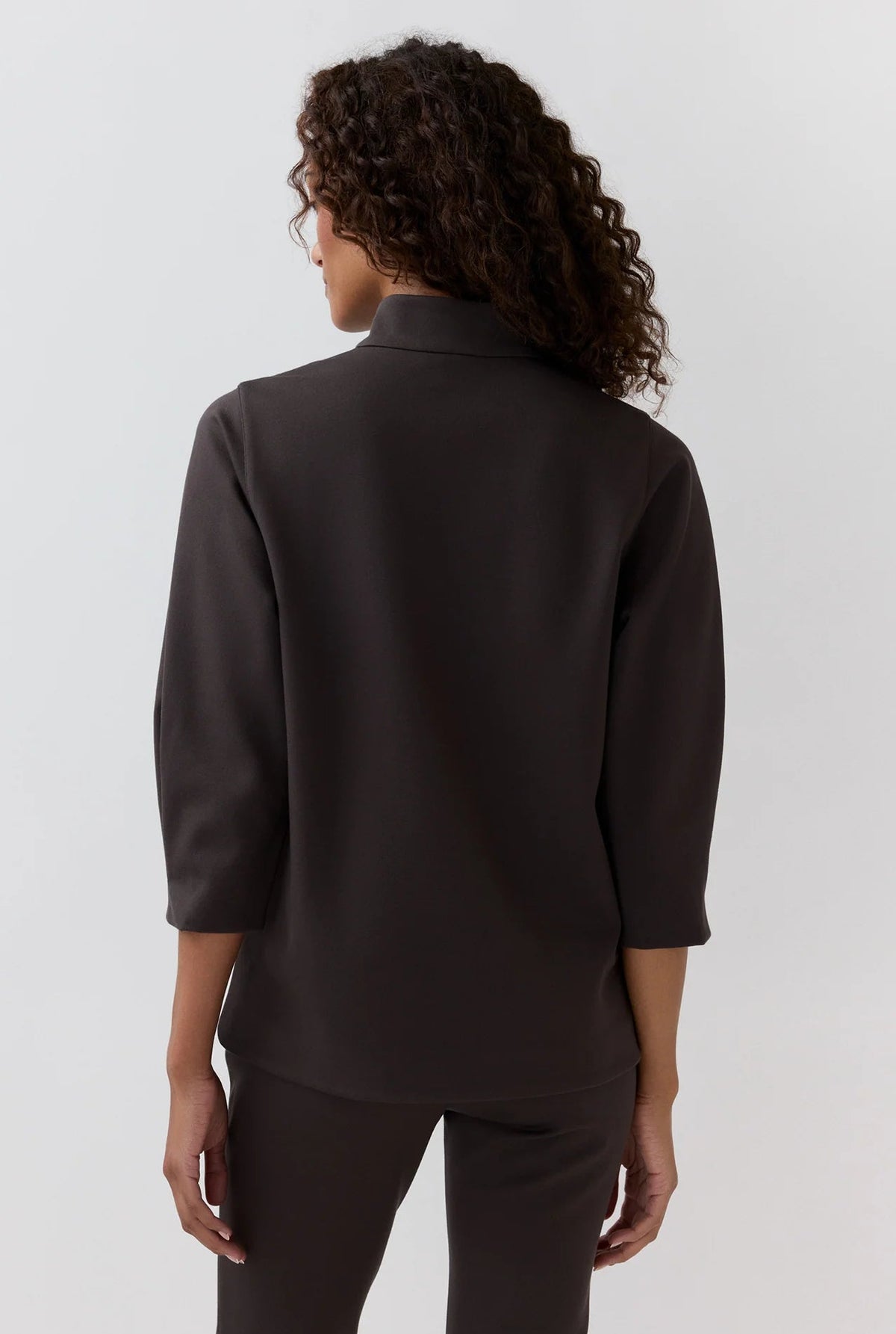 A person with curly hair stands with their back to the camera against a plain light background, wearing the Ann Mashburn Violetta Bi-Stretch Tricotine Tunic—a modern, dark, long-sleeve, stretchy top—paired with matching pants.