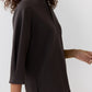 A person wearing the Ann Mashburn Violetta Bi-Stretch Tricotine Tunic, a loose-fitting, long-sleeve women’s top with a high collar, photographed from the side against a plain background.