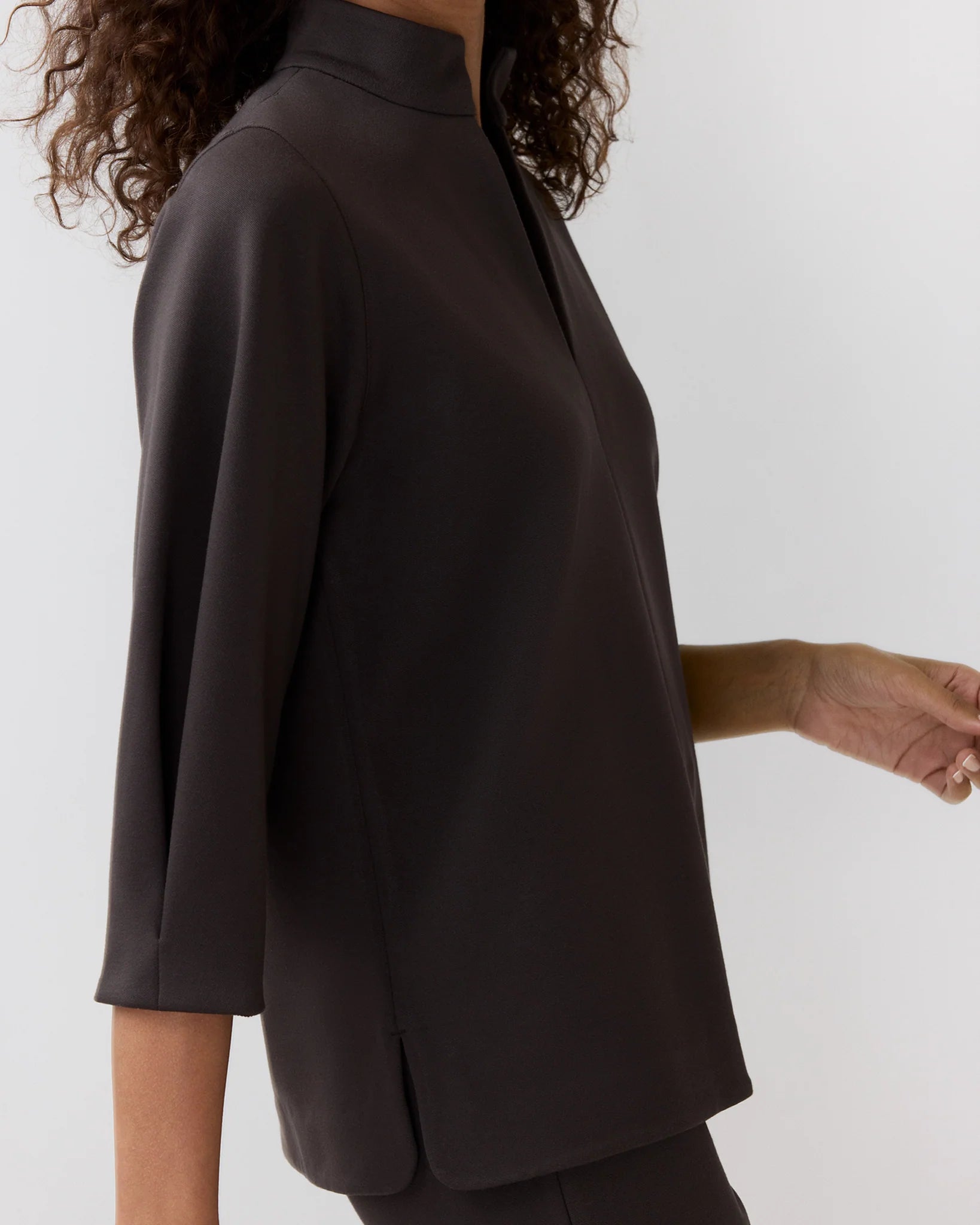 A person wearing the Ann Mashburn Violetta Bi-Stretch Tricotine Tunic, a loose-fitting, long-sleeve women’s top with a high collar, photographed from the side against a plain background.