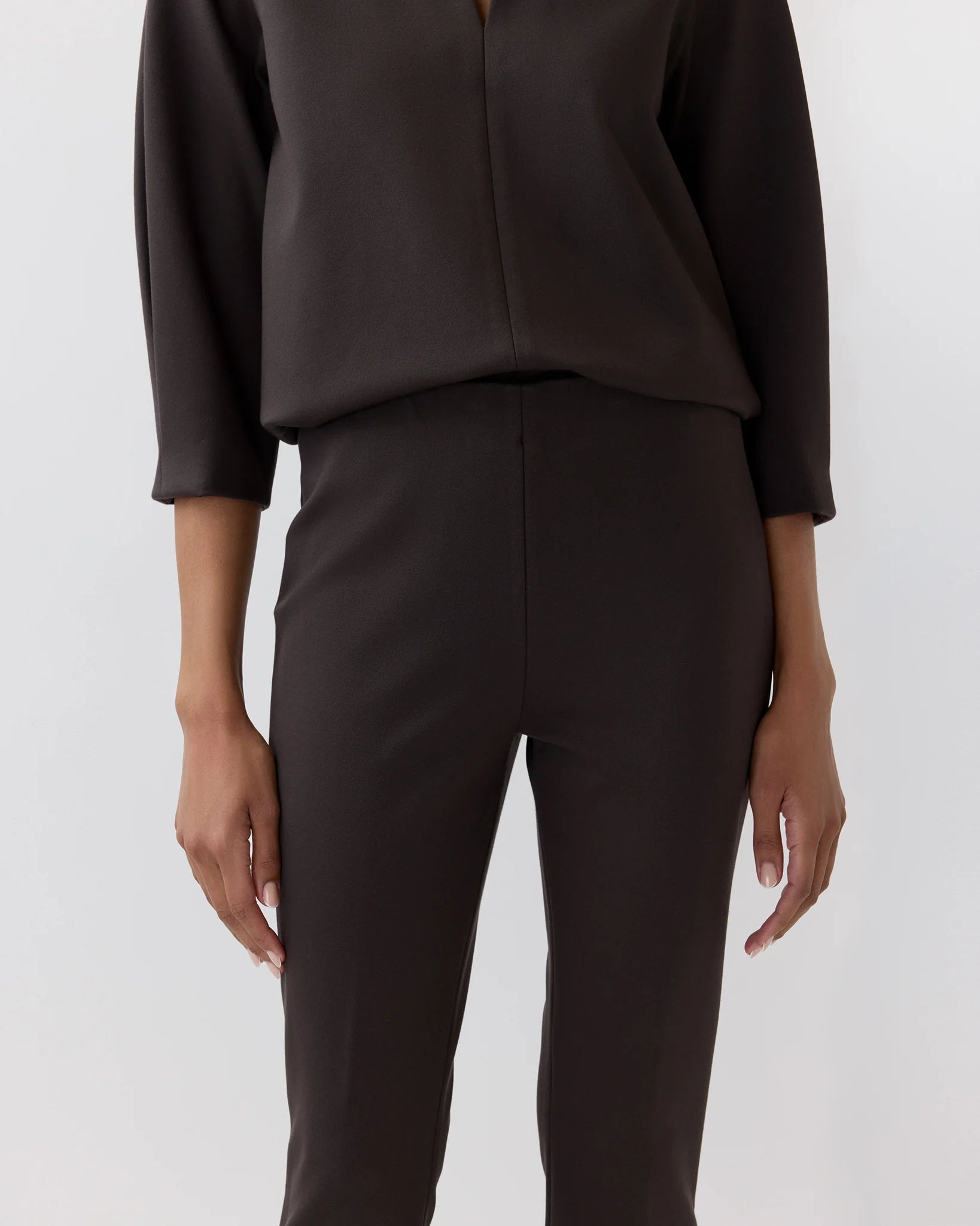 A person wearing Ann Mashburn’s Faye Flare Cropped Pant in dark brown with a matching long-sleeve blouse, standing against a plain light background.