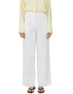 A person wearing Callas Milano Sailor Pants in white, paired with a pale yellow button-up shirt and black sandals, stands against a white background.