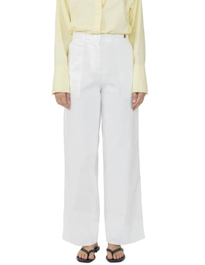A person wearing Callas Milano Sailor Pants in white, paired with a pale yellow button-up shirt and black sandals, stands against a white background.