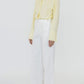 A person wearing a pale yellow long-sleeve shirt with decorative collar accents, Callas Milano Sailor Pant in white high-waisted relaxed wide-leg fit, and black sandals stands against a plain background.