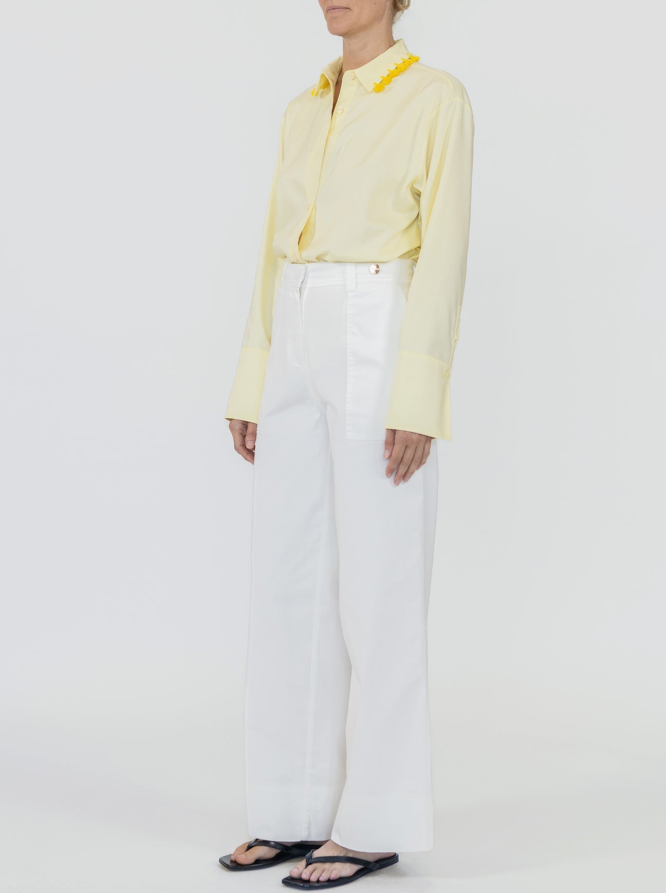 A person wearing a pale yellow long-sleeve shirt with decorative collar accents, Callas Milano Sailor Pant in white high-waisted relaxed wide-leg fit, and black sandals stands against a plain background.