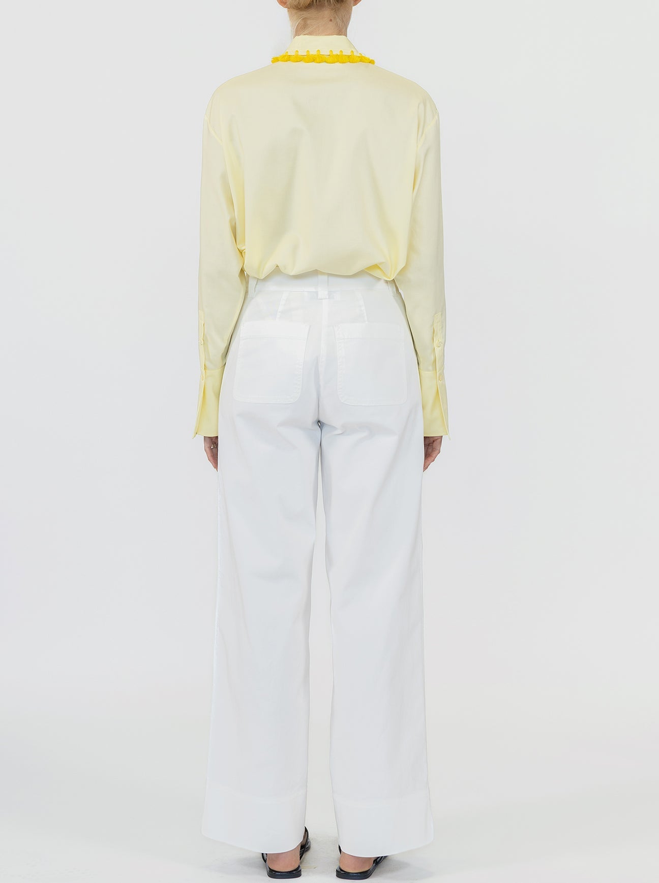 A person stands facing away, wearing Callas Milano’s Sailor Pant—a high-waisted, white wide leg pant in soft cotton twill—paired with a light yellow long-sleeve shirt against a plain white background.