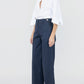 A person stands against a plain background wearing Callas Milano's Sailor Pant—a navy high-waisted cotton twill wide-leg style—paired with a white button-up shirt with rolled sleeves and black sandals.