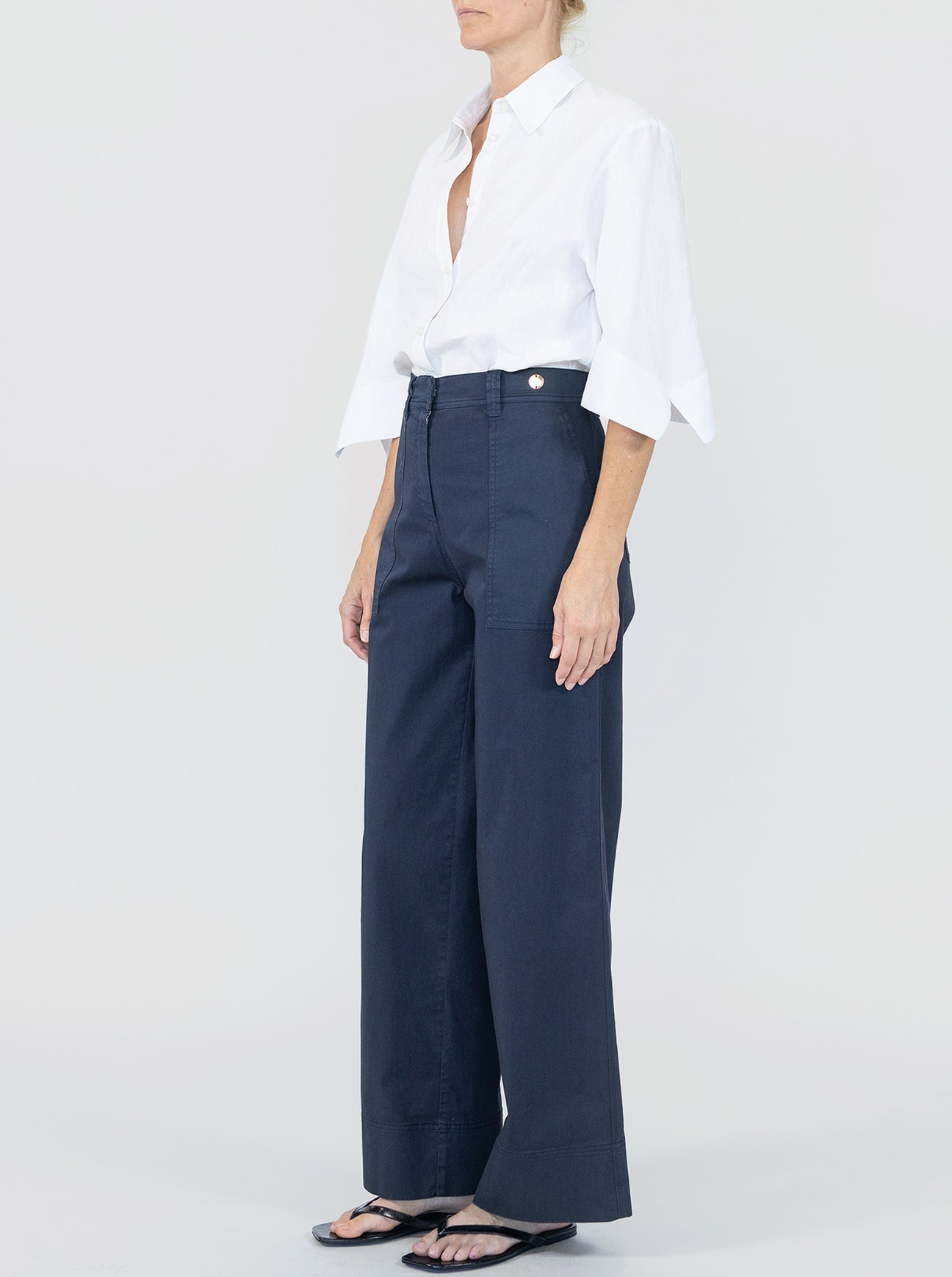 A person stands against a plain background wearing Callas Milano's Sailor Pant—a navy high-waisted cotton twill wide-leg style—paired with a white button-up shirt with rolled sleeves and black sandals.