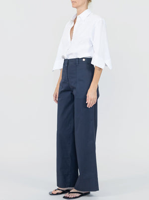 A person stands against a plain background wearing Callas Milano's Sailor Pant—a navy high-waisted cotton twill wide-leg style—paired with a white button-up shirt with rolled sleeves and black sandals.