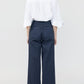 A person faces away, wearing a white collared shirt with elbow-length sleeves and Callas Milano Sailor Pant—navy blue, high-waisted, relaxed fit, wide leg cotton twill—against a plain light background.