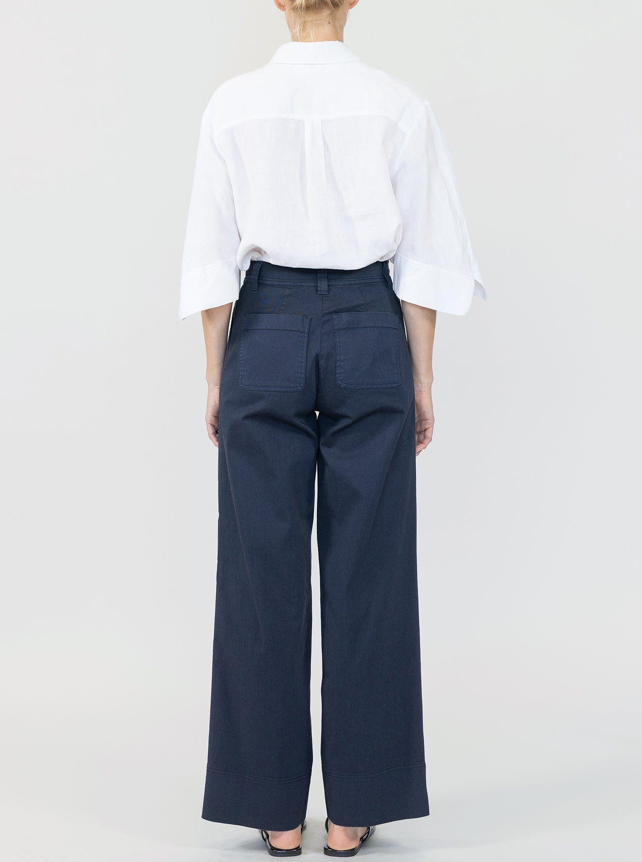 A person faces away, wearing a white collared shirt with elbow-length sleeves and Callas Milano Sailor Pant—navy blue, high-waisted, relaxed fit, wide leg cotton twill—against a plain light background.