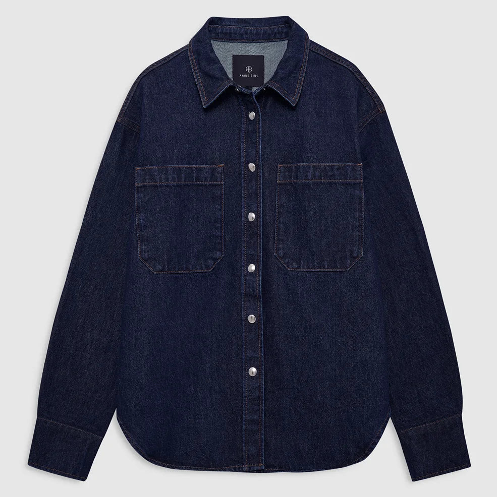 The Samy Shirt by Anine Bing, crafted from dark blue cotton denim canvas, features silver buttons, two spacious chest pockets, and a pointed collar; it is displayed laid flat on a plain light background.
