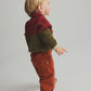 A toddler stands on a light-colored floor, wearing a red and green sweater, beige shoes, and rust-colored Cozmo Baby Saul Twill Pants, facing slightly away from the camera.