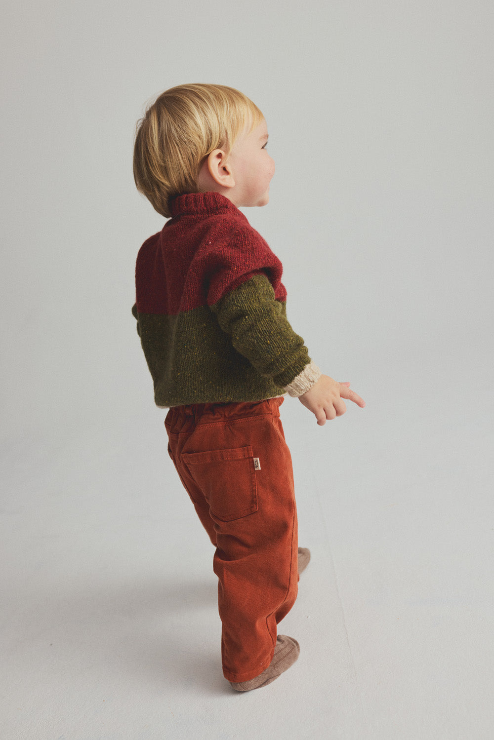 A toddler stands on a light-colored floor, wearing a red and green sweater, beige shoes, and rust-colored Cozmo Baby Saul Twill Pants, facing slightly away from the camera.