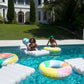 Two people are enjoying a sunny day in a pool with large inflatable rafts and a Sunnylife Beach Ball, Pool Side Pastel Gelato from Sunnylife, in front of a white mansion with a manicured lawn—true summer fun.