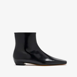 The Proenza Schouler Trap Flat Ankle Boots feature black nappa leather with a pointed toe, low square heel, and glossy finish, shown from the side against a plain white background.