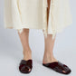 A person wearing a cream-colored textured skirt and dark burgundy Proenza Schouler Slant Cross Slides stands on a light gray floor.