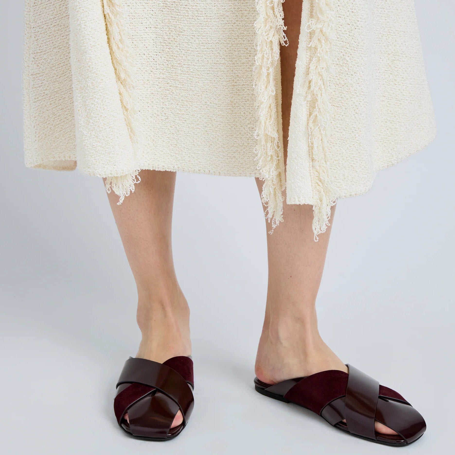 A person wearing a cream-colored textured skirt and dark burgundy Proenza Schouler Slant Cross Slides stands on a light gray floor.