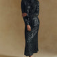 A woman stands against a neutral beige background, wearing the Sequin Lucy Shirt by Lela Rose—a long-sleeve, black sequined collared top that makes a true statement—paired with black pointed shoes.