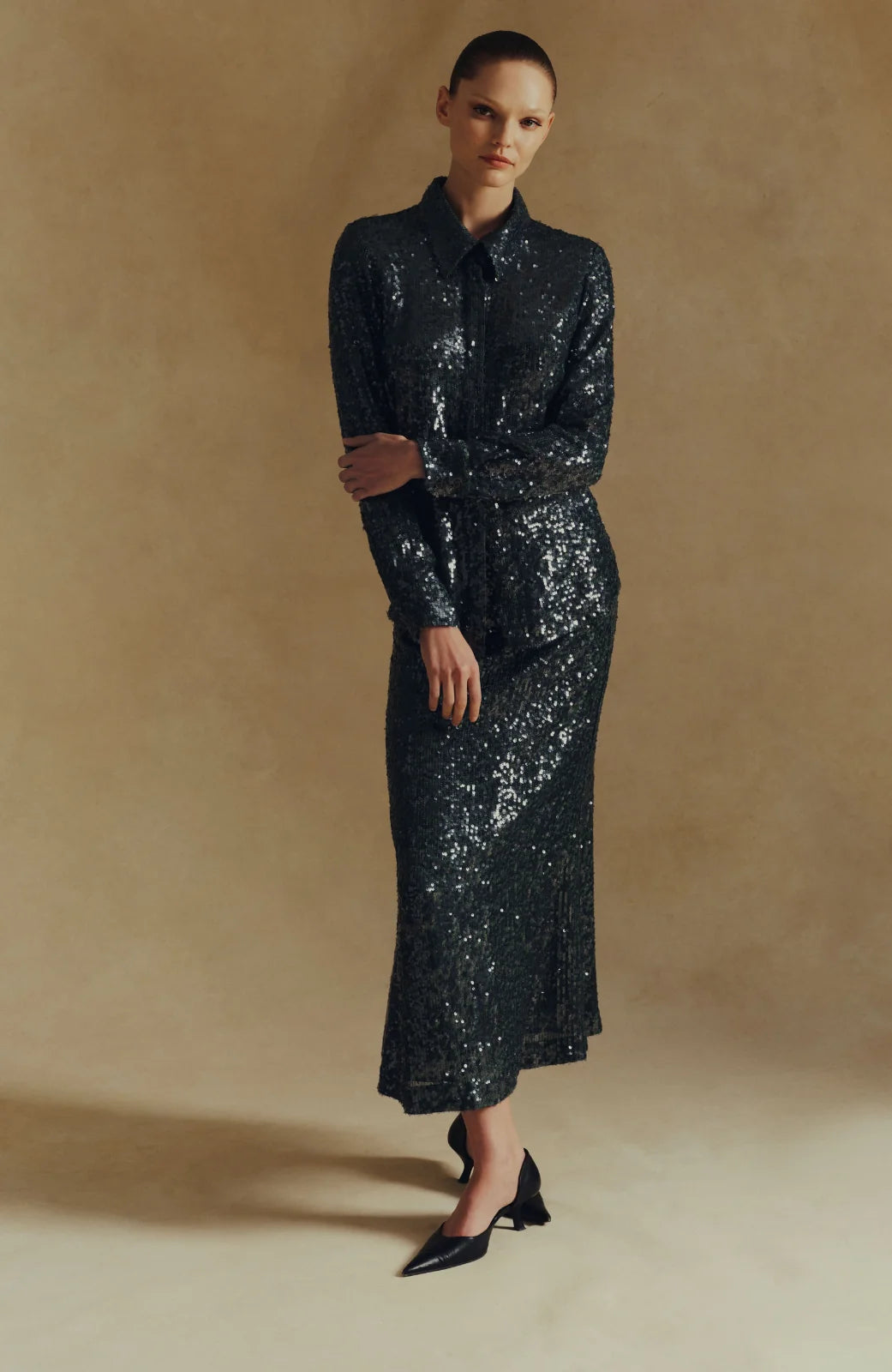 A woman stands against a neutral beige background, wearing the Sequin Lucy Shirt by Lela Rose—a long-sleeve, black sequined collared top that makes a true statement—paired with black pointed shoes.