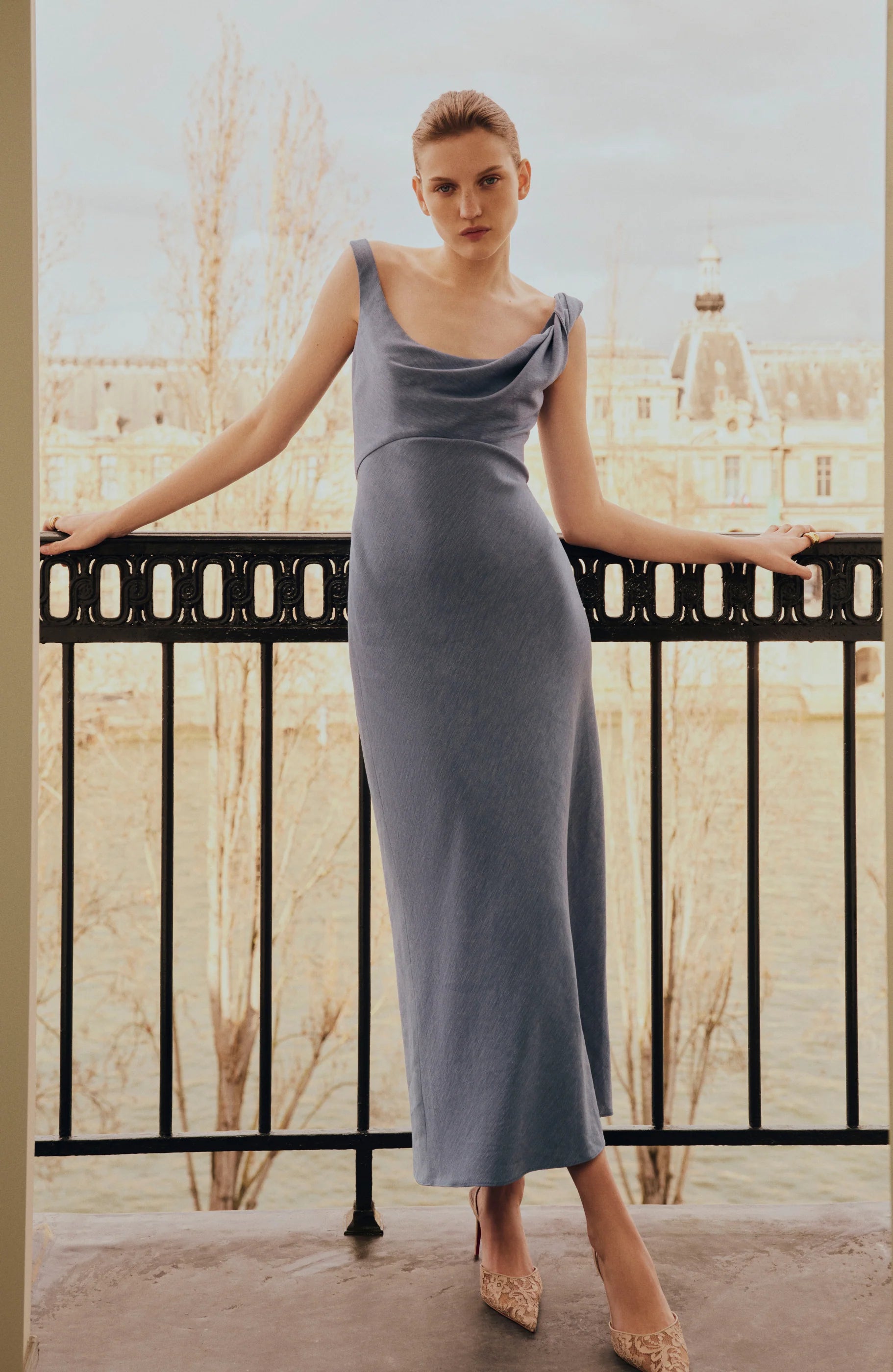 A woman stands on a balcony overlooking a river and historic buildings, wearing the Lela Rose Linen Twist Detail Dress—a sleeveless, light blue, anti-wrinkle linen piece with an A-line skirt.