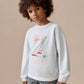A child with curly hair stands against a beige background, wearing the Bonpoint Boys' Erol Embroidered Regatta Sweatshirt and striped pants.
