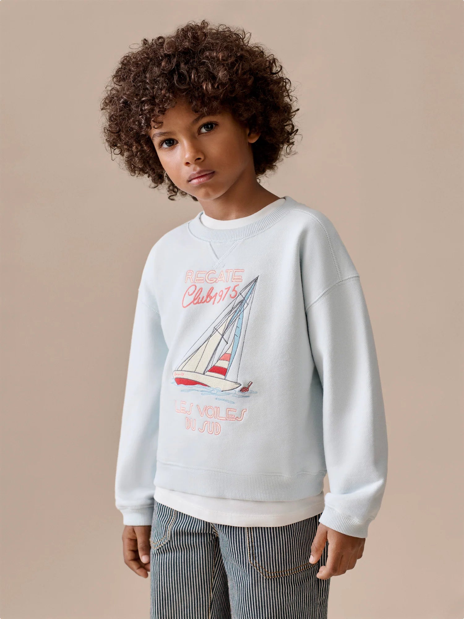 A child with curly hair stands against a beige background, wearing the Bonpoint Boys' Erol Embroidered Regatta Sweatshirt and striped pants.
