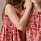 Two young girls in Bonpoint Girls' Goldia blouses in Liberty Fabric stand close; one whispers into the other's ear while gently touching her face.