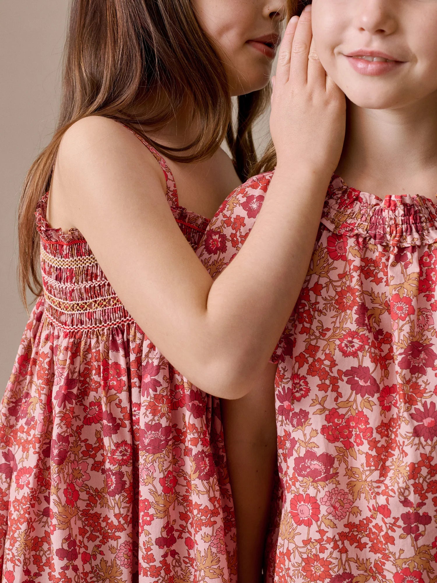Two young girls in Bonpoint Girls' Goldia blouses in Liberty Fabric stand close; one whispers into the other's ear while gently touching her face.