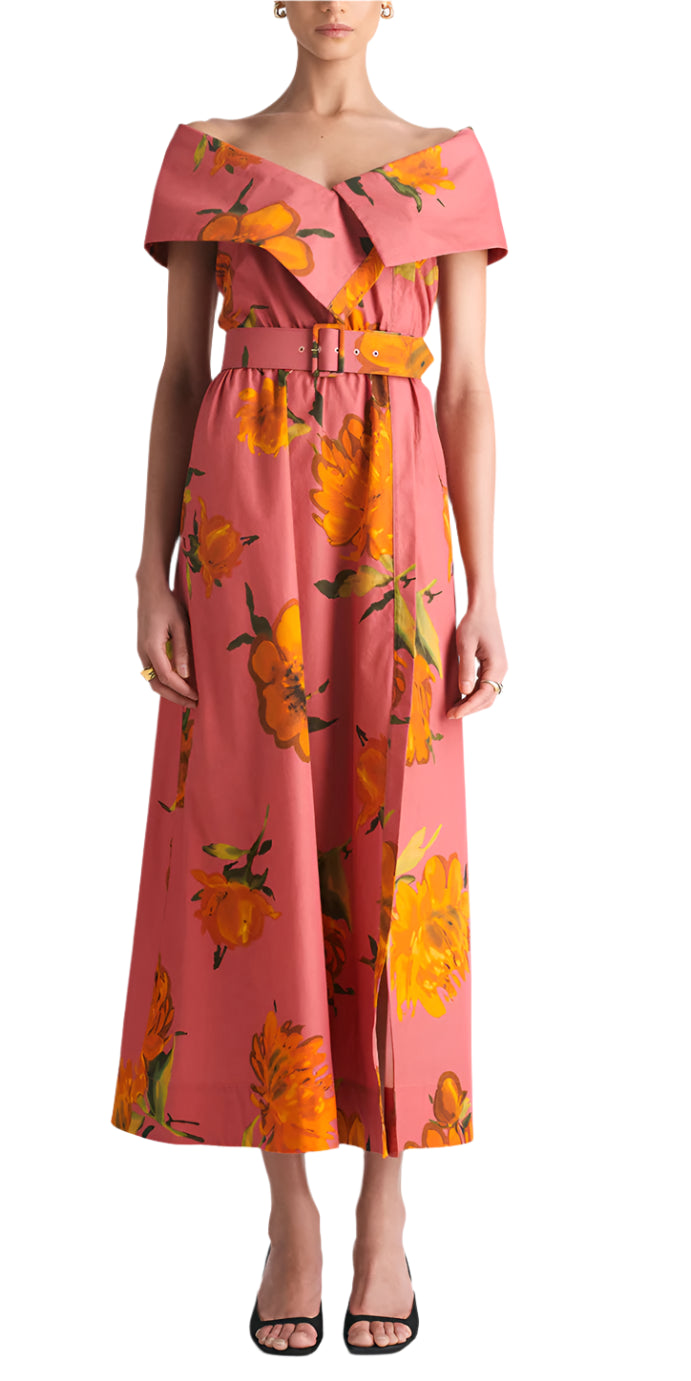 A woman models the Lela Rose Asymmetrical Cuff Dress—pink, off-shoulder with yellow floral print and cinched waist—paired with black open-toe heels, standing against a plain white background.