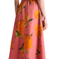 A woman in a Lela Rose Asymmetrical Cuff Dress with a pink, yellow-orange floral print and cinched waist, paired with black sandals, is shown from the back.