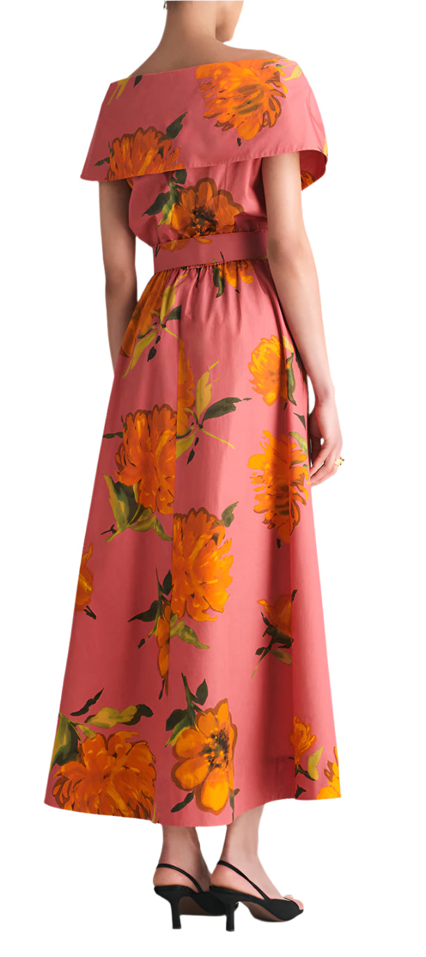 A woman in a Lela Rose Asymmetrical Cuff Dress with a pink, yellow-orange floral print and cinched waist, paired with black sandals, is shown from the back.