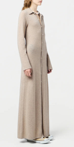 A person stands in profile wearing the Guest in Residence Showtime Cashmere Shirt Dress, a lightweight beige button-down with flared sleeves, paired with white sneakers against a plain background.